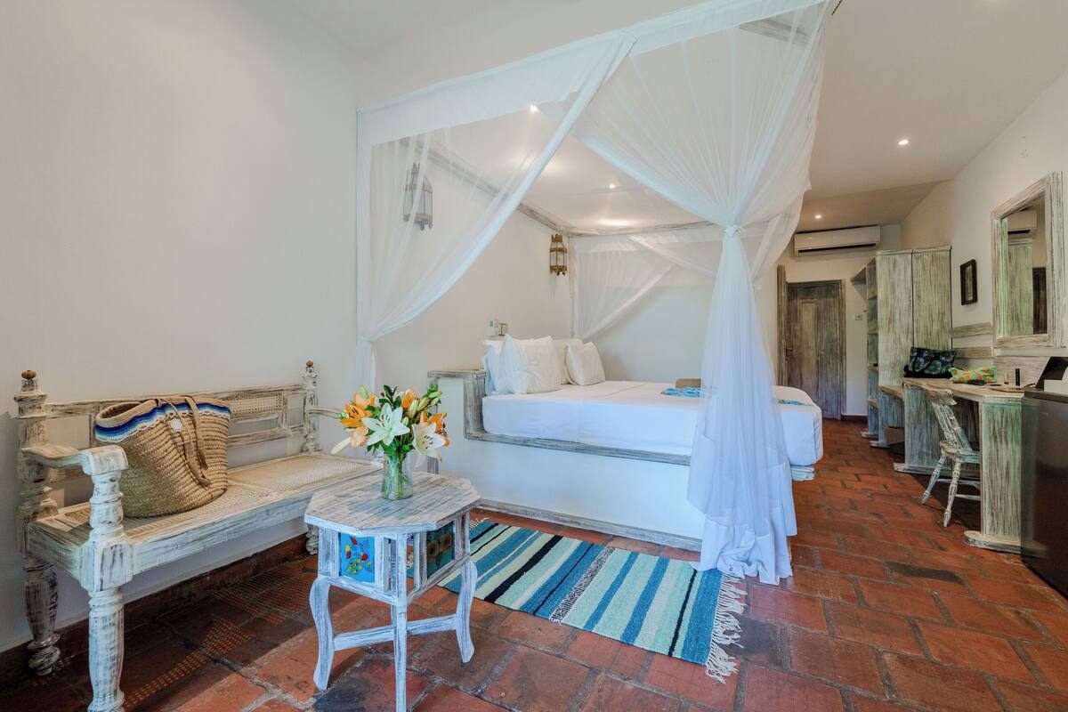 A bedroom with a four-poster bed, a bench with a woven bag, a small table with a vase of flowers, and a striped rug.
