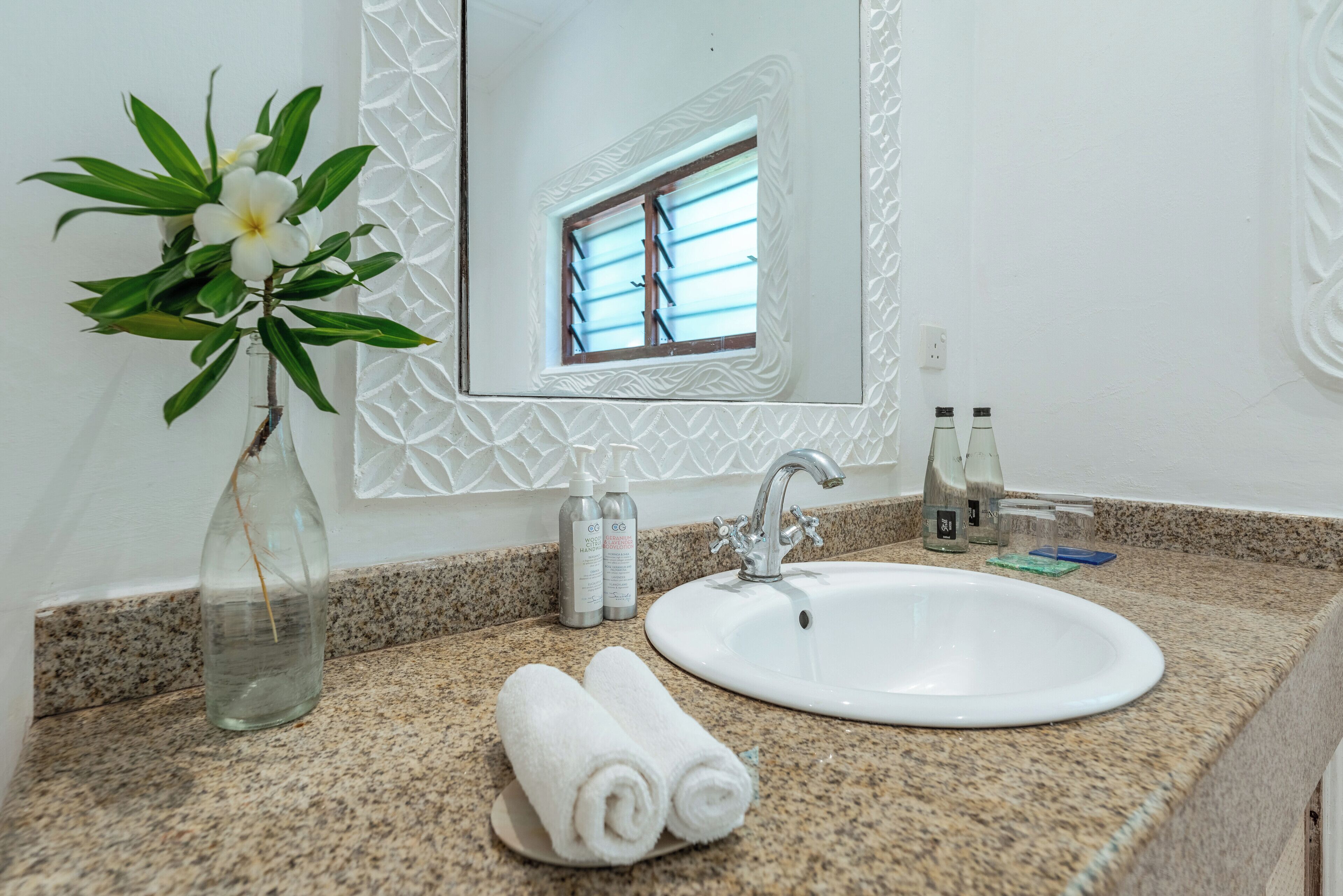 Un baño con encimera de granito, un lavamanos blanco, un espejo con marco decorativo, un florero con una flor y artículos de tocador.