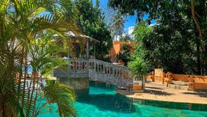Una piscina cristalina con un puente, rodeada de exuberante vegetación y una zona sombreada con una pérgola.