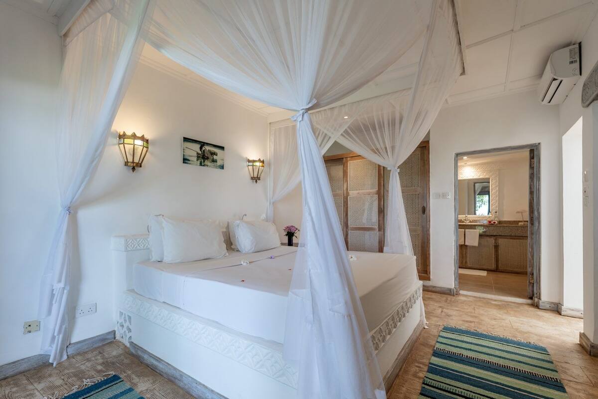 A bedroom with a four-poster bed, a bedside table, and a nightstand. There is a window with a view of the outside, and a door leading to the bathroom.