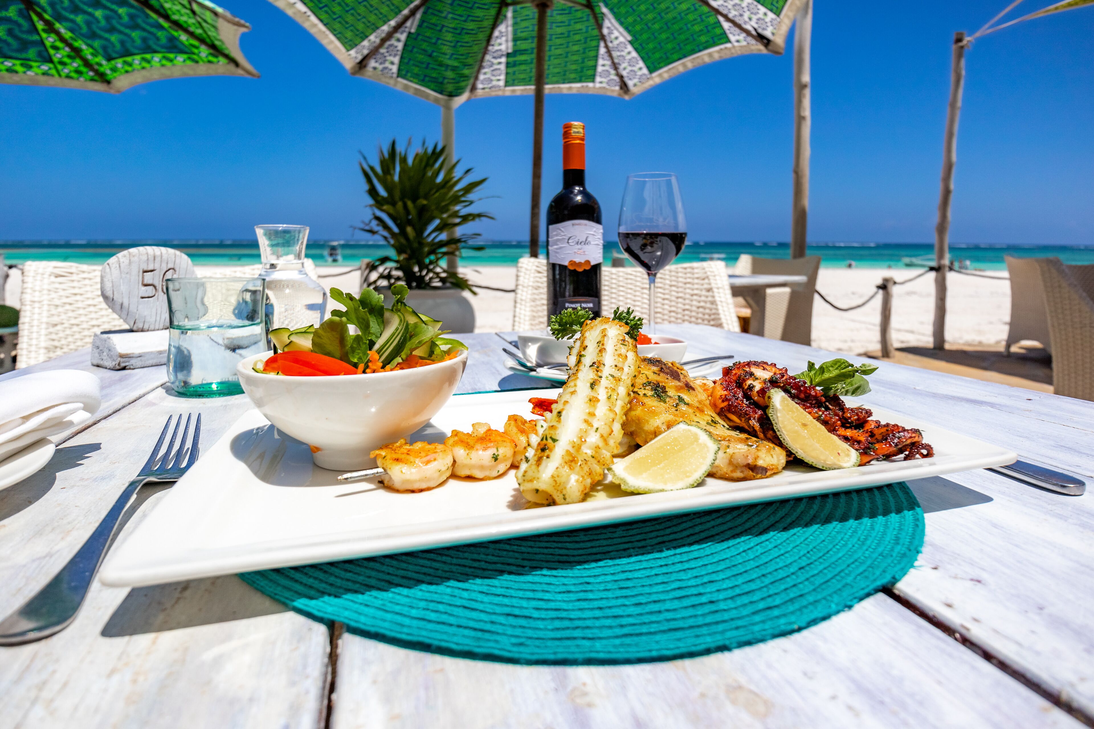A table set with seafood, a wine bottle, and a glass of red wine on a beach with umbrellas and chairs.