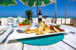 A table set with seafood, a wine bottle, and a glass of red wine on a beach with umbrellas and chairs.