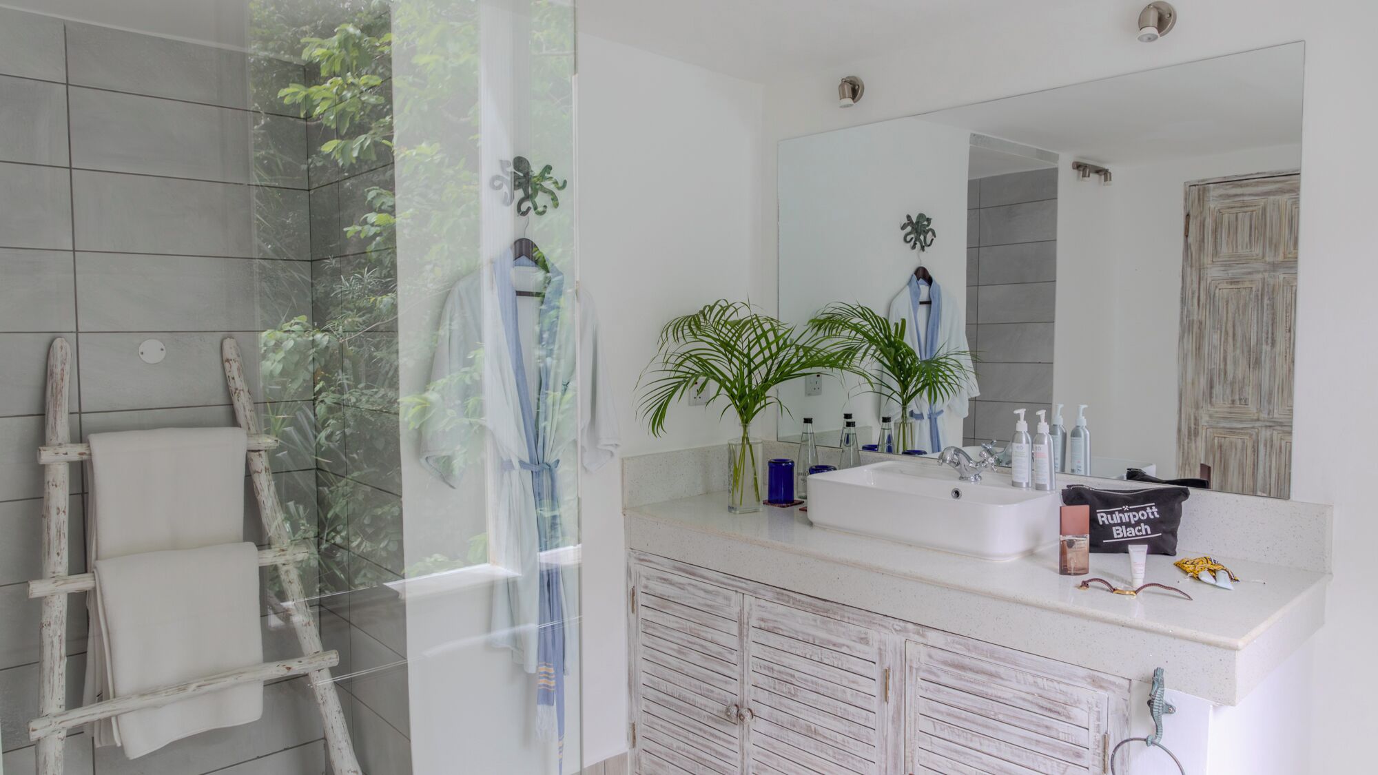 Un baño con un espejo grande, dos lavamanos, un perchero para toallas y una zona de ducha.
