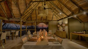 A dining area with a long table set for a meal, wooden beams on the ceiling, and a view of trees outside. - Ghoha Hills Savuti (Chobe National Park)
