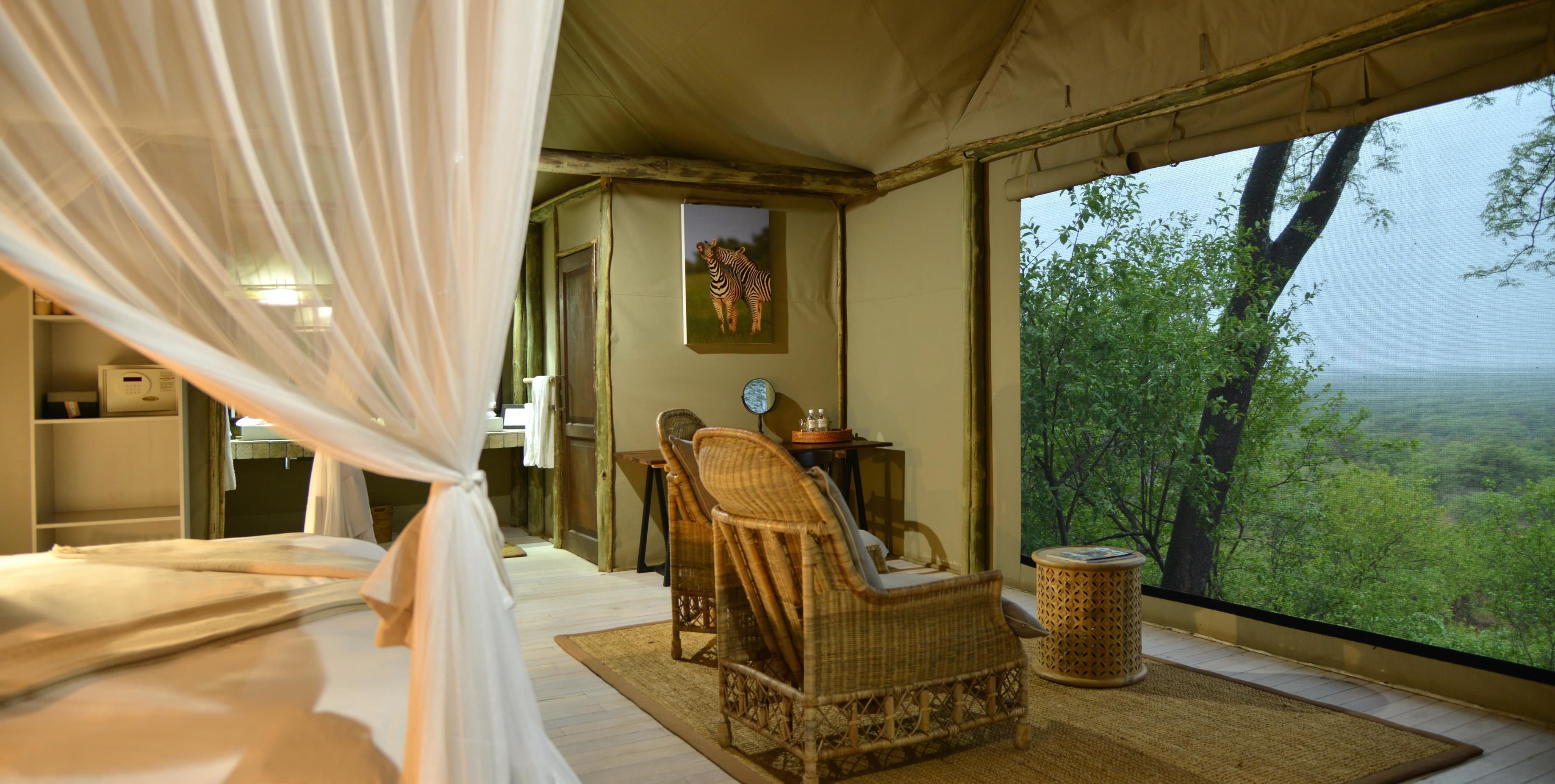Una habitación con carpa, una cama, una silla de mimbre, una mesita y vista al bosque.