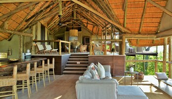 Interior rústico com telhado de palha, móveis de vime e uma escada que leva a uma varanda.