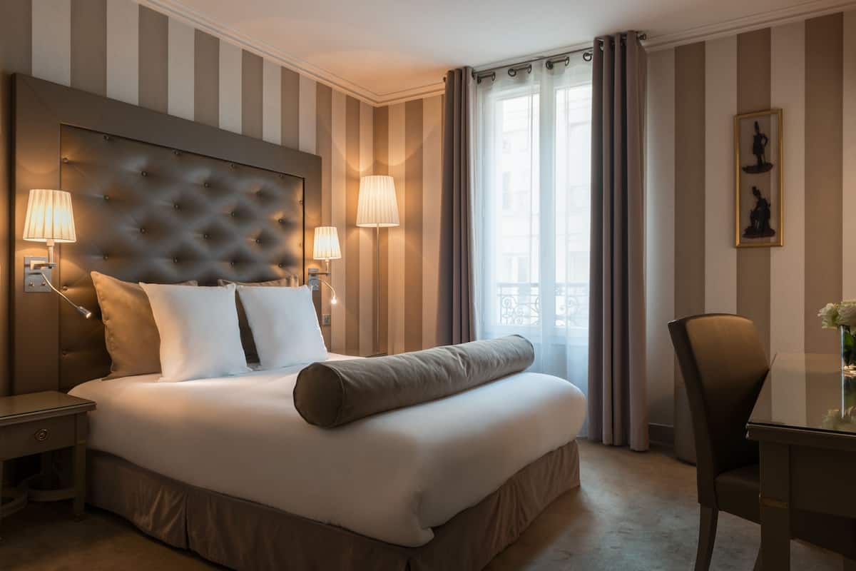 A hotel room with a large bed, two bedside lamps, a headboard, a window with curtains, and a framed picture on the wall.