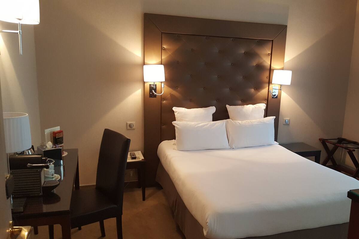 A hotel room with a large bed, two bedside lamps, a desk with a chair, and a small table.