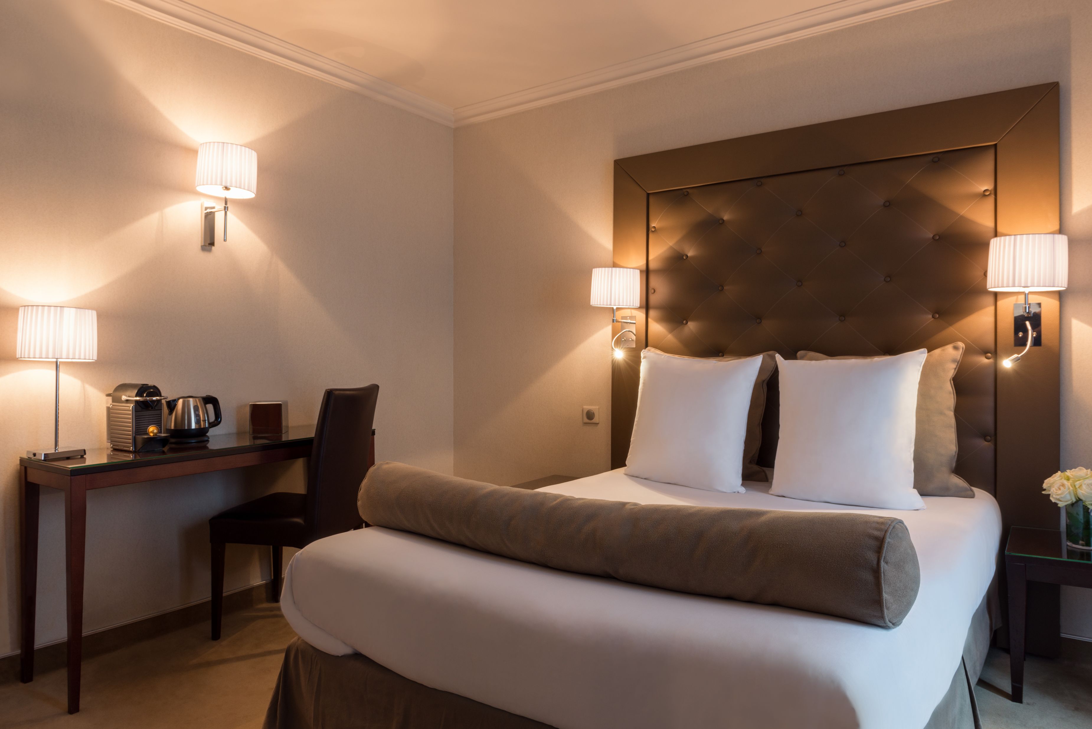 A hotel room with a large bed, two bedside lamps, a desk with a coffee maker, and a chair.