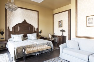 A double bed with a wooden headboard, a bench, a sofa, a chandelier, and framed pictures on the walls.