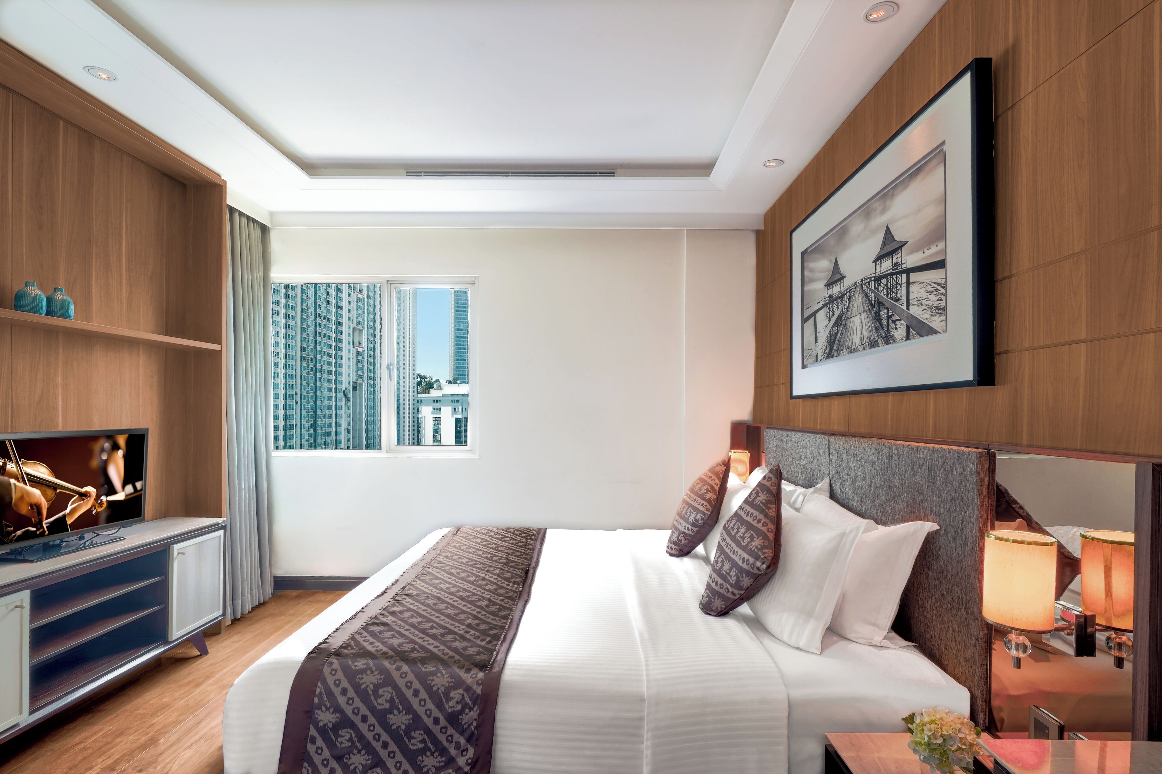 Une chambre d’hôtel moderne avec un grand lit, une télévision, une armoire en bois, une fenêtre donnant sur la ville et un tableau encadré au mur.