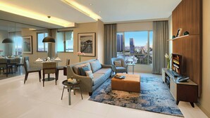 Salón moderno de hotel con sofá, sillón, mesa de comedor y televisor sobre un mueble de madera.