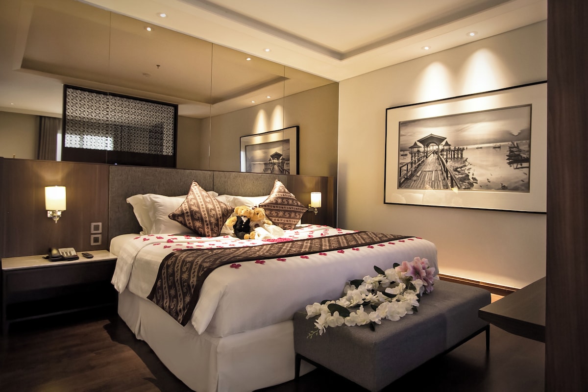 A hotel room with a large bed, bedside lamps, a nightstand, a desk, and a framed picture on the wall.