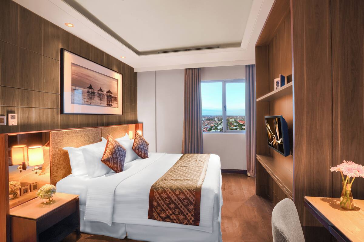 A hotel room with a large bed, bedside lamps, a flat-screen TV, a view of the city, and a wooden wall panel.