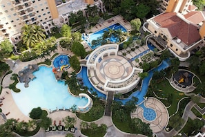 Vista aérea de um resort com várias piscinas, palmeiras e um espelho d'água circular central.