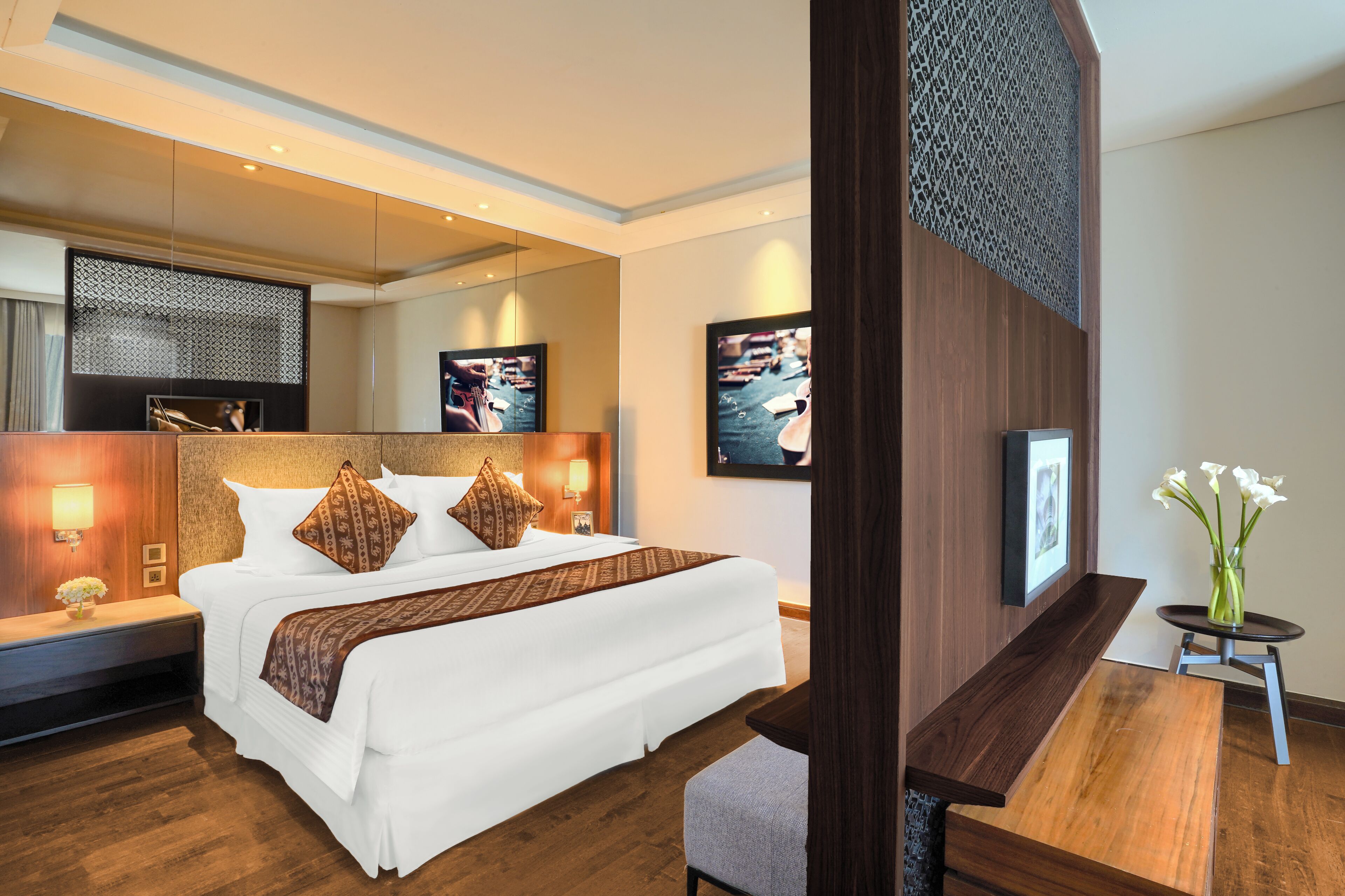 A hotel room with a large bed, bedside tables, a small round table, a chair, a vase with flowers, and wall-mounted artwork.