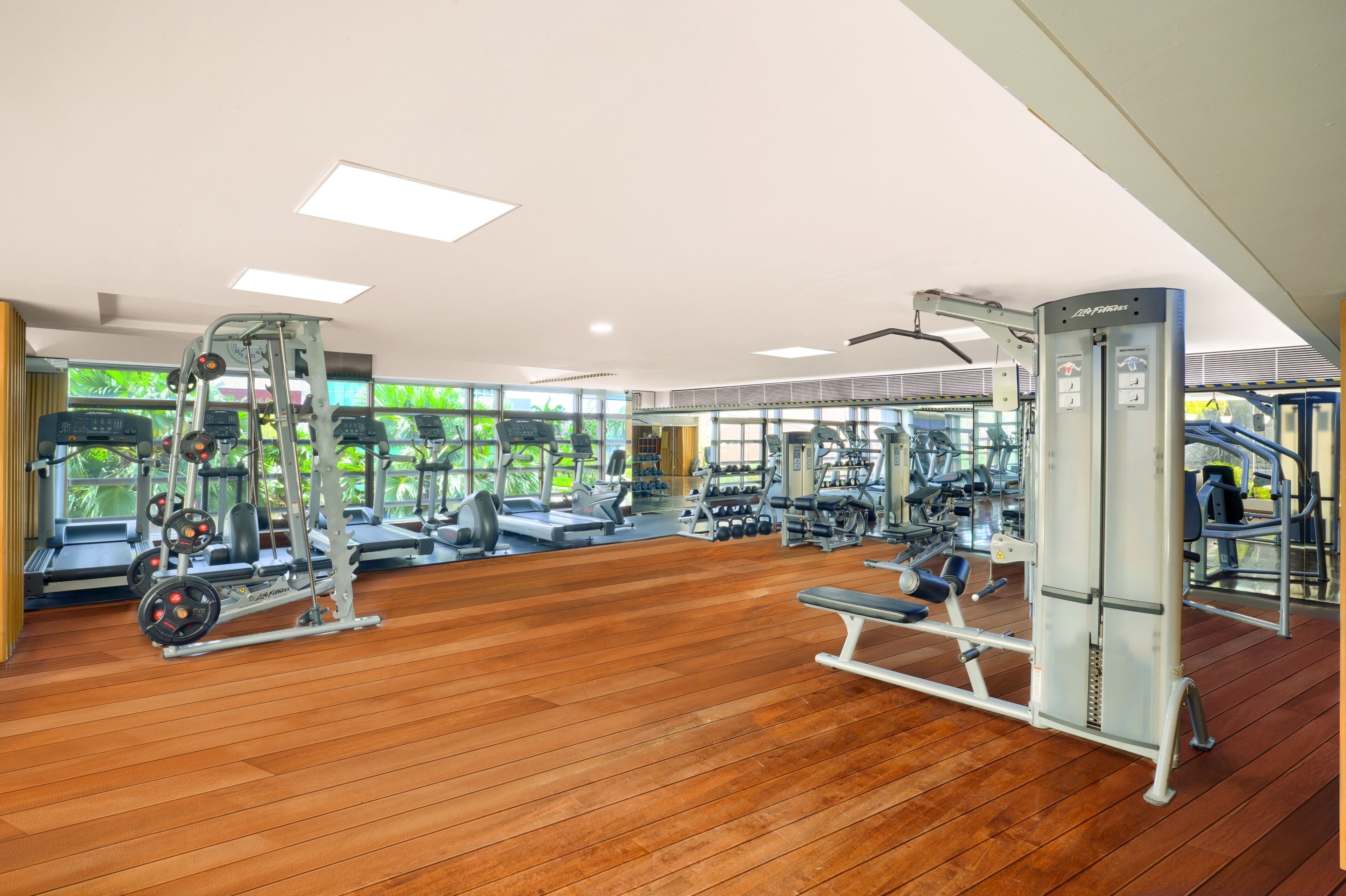 A gym with various exercise equipment, wooden flooring, and large windows with a view of greenery.