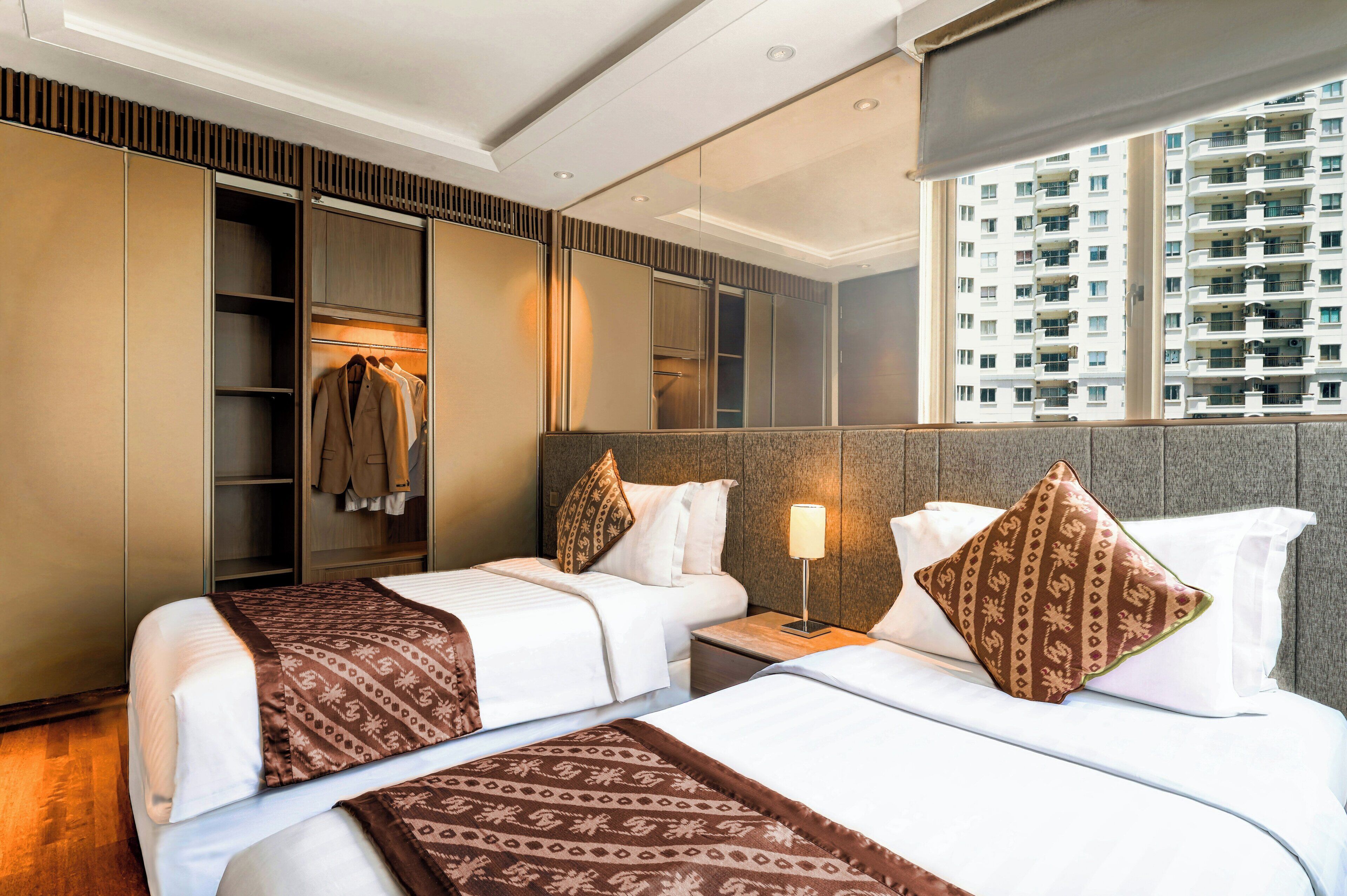 A hotel room with two beds, a wardrobe, a mirror, a nightstand, and a window with a view of a high-rise building.