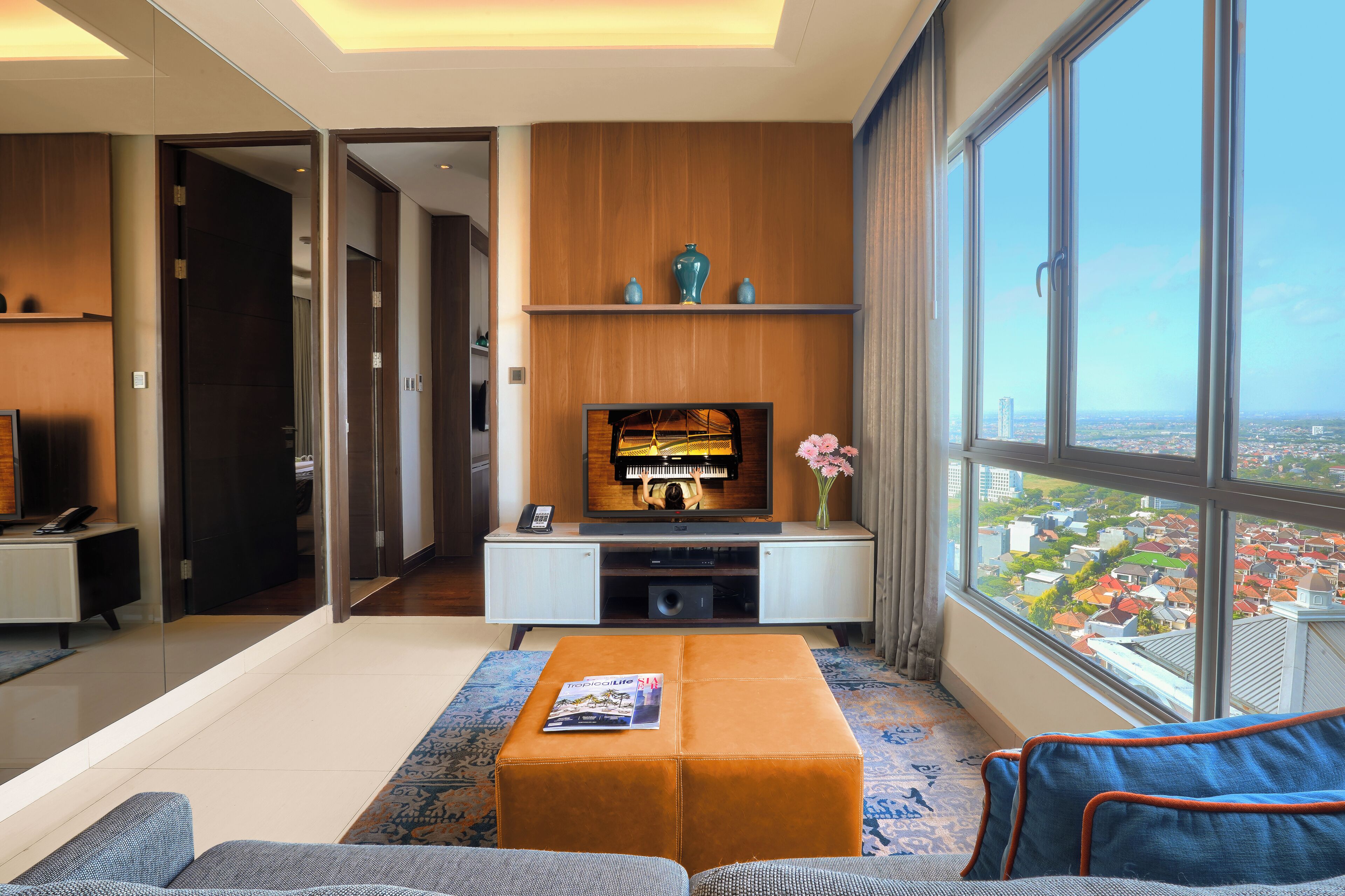 A modern living room with a television, sofa, ottoman, and a large window offering a city view.