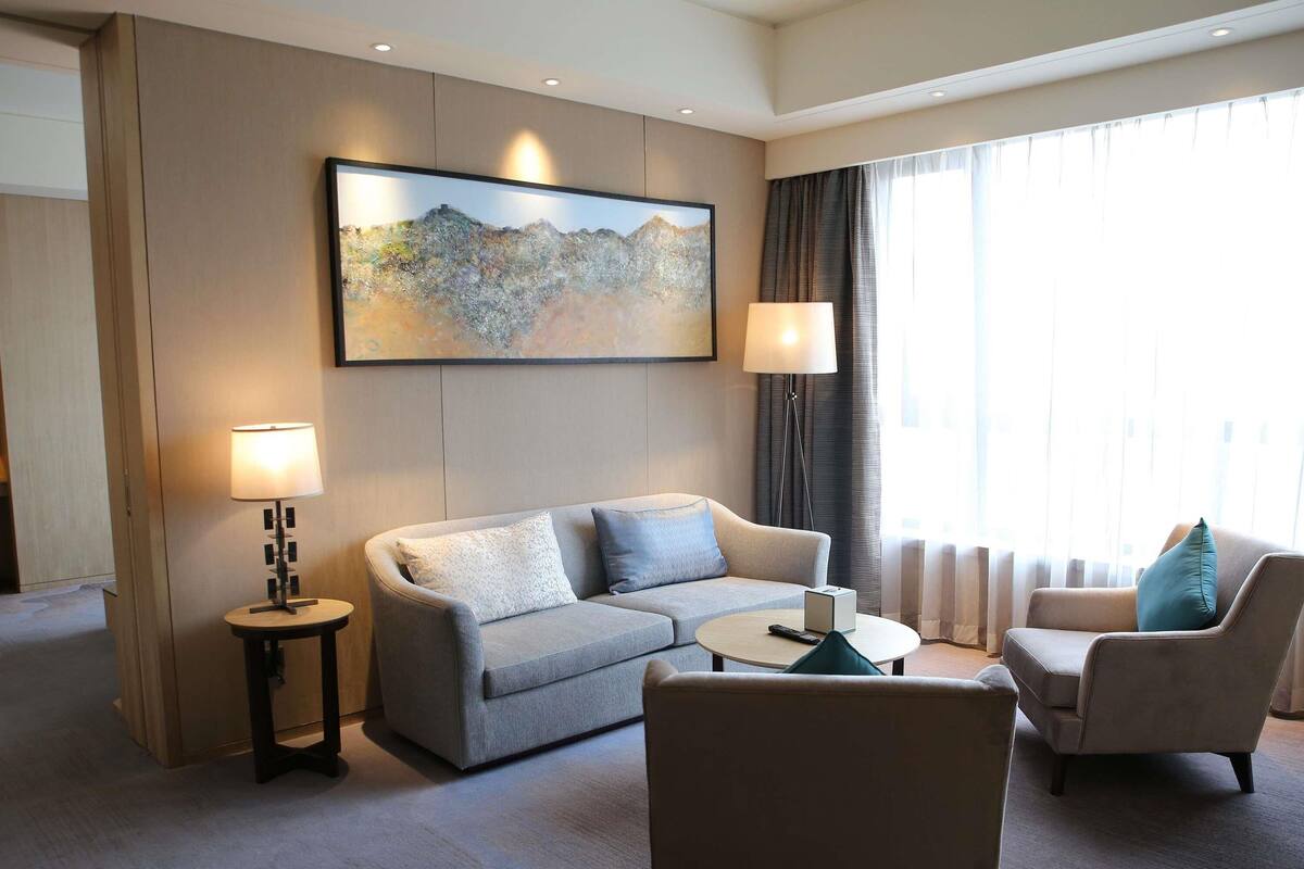A hotel room with a sofa, armchair, a small table, a lamp, and a painting on the wall.