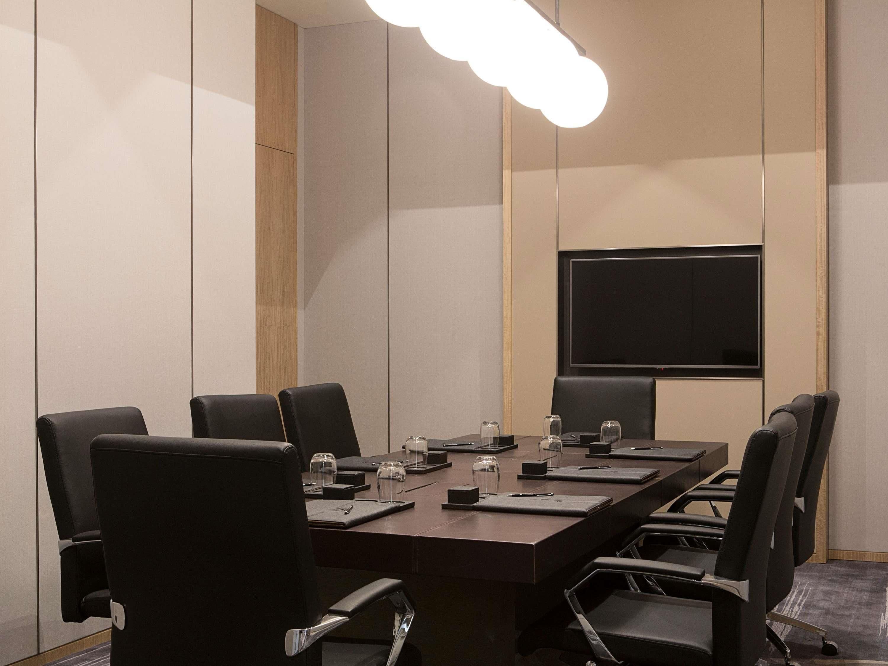 Uma sala de conferências com uma mesa comprida, cadeiras pretas, uma TV de tela plana e um lustre moderno.
