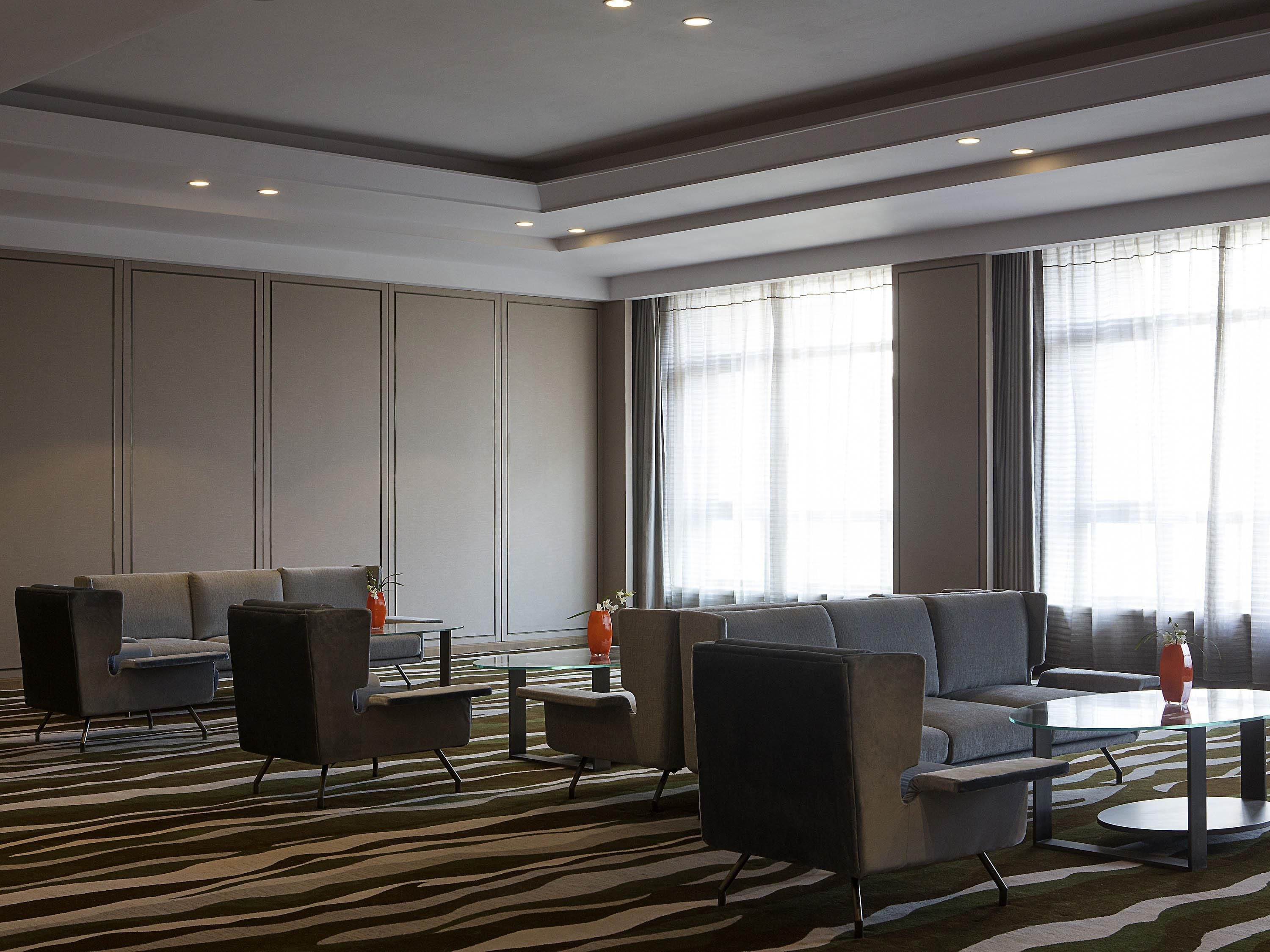 A modern hotel lobby with a zebra-patterned carpet, a sofa, armchairs, a glass coffee table, and large windows with sheer curtains.