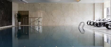 An indoor swimming pool with clear water, a marble wall, and a seating area with lounge chairs.