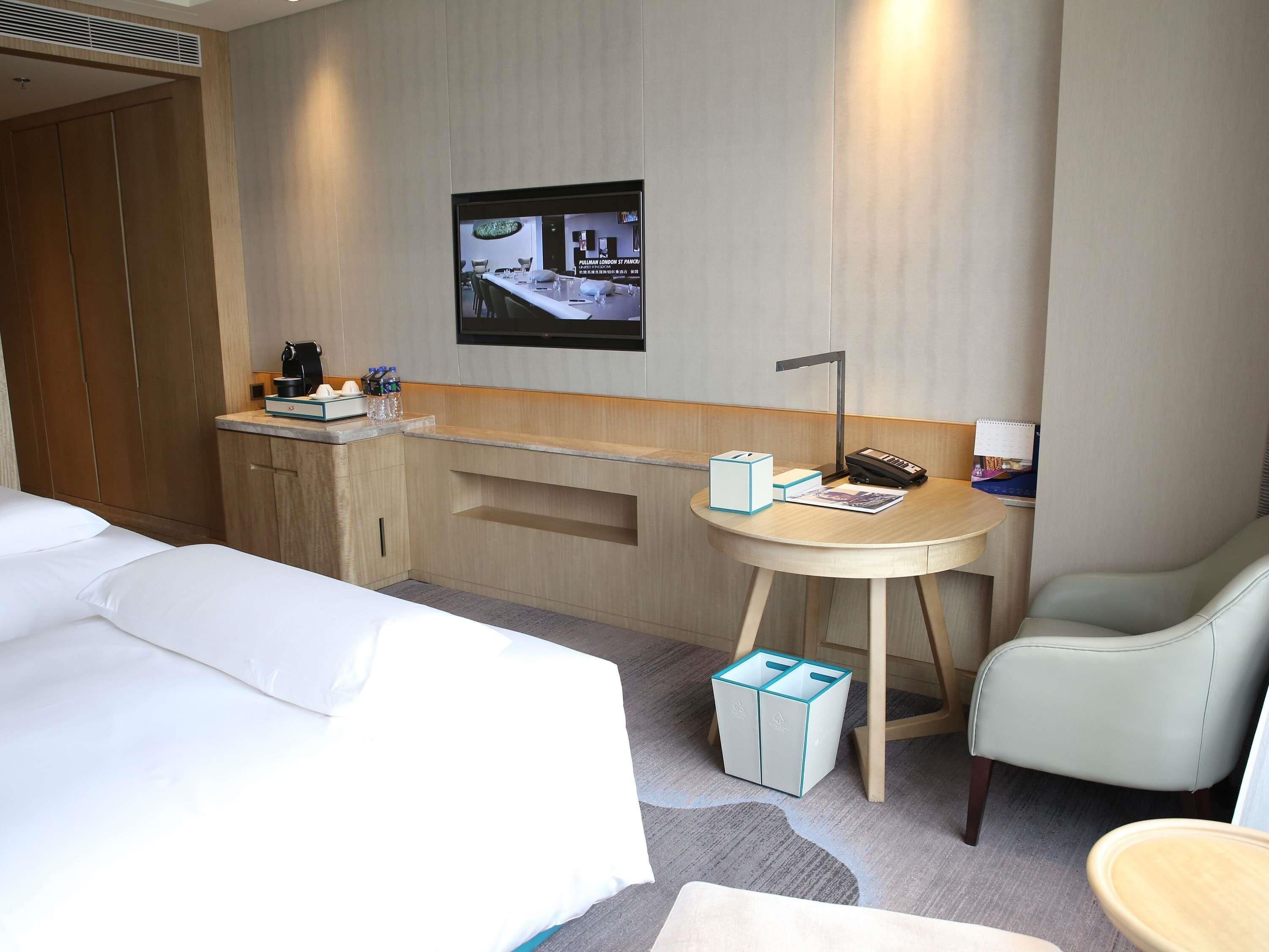 A hotel room with a bed, a desk with a chair, a small table, a trash bin, and a wall-mounted TV.
