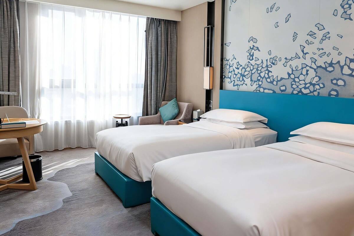 A hotel room with two beds, a desk, a chair, a trash bin, and a large window with curtains.