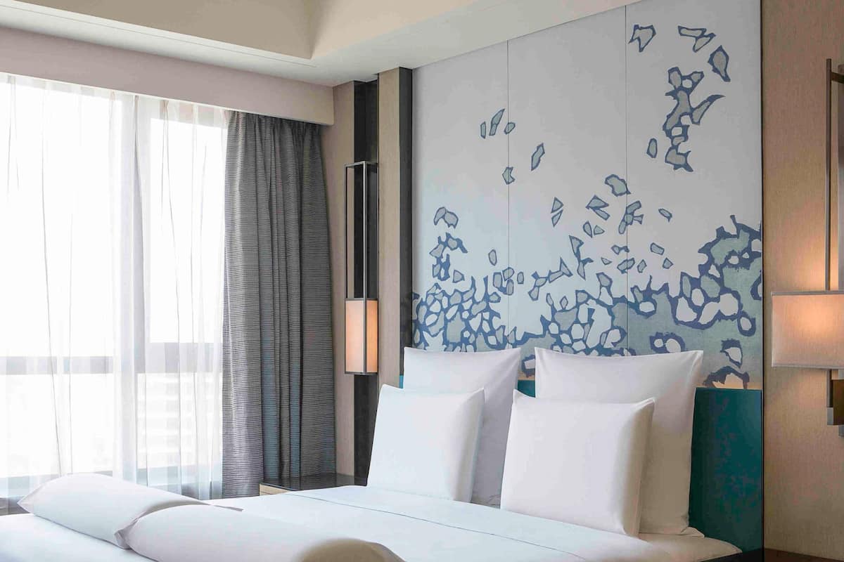 A hotel room with a bed, curtains, a wall with a blue abstract pattern, and a bedside table with a lamp.