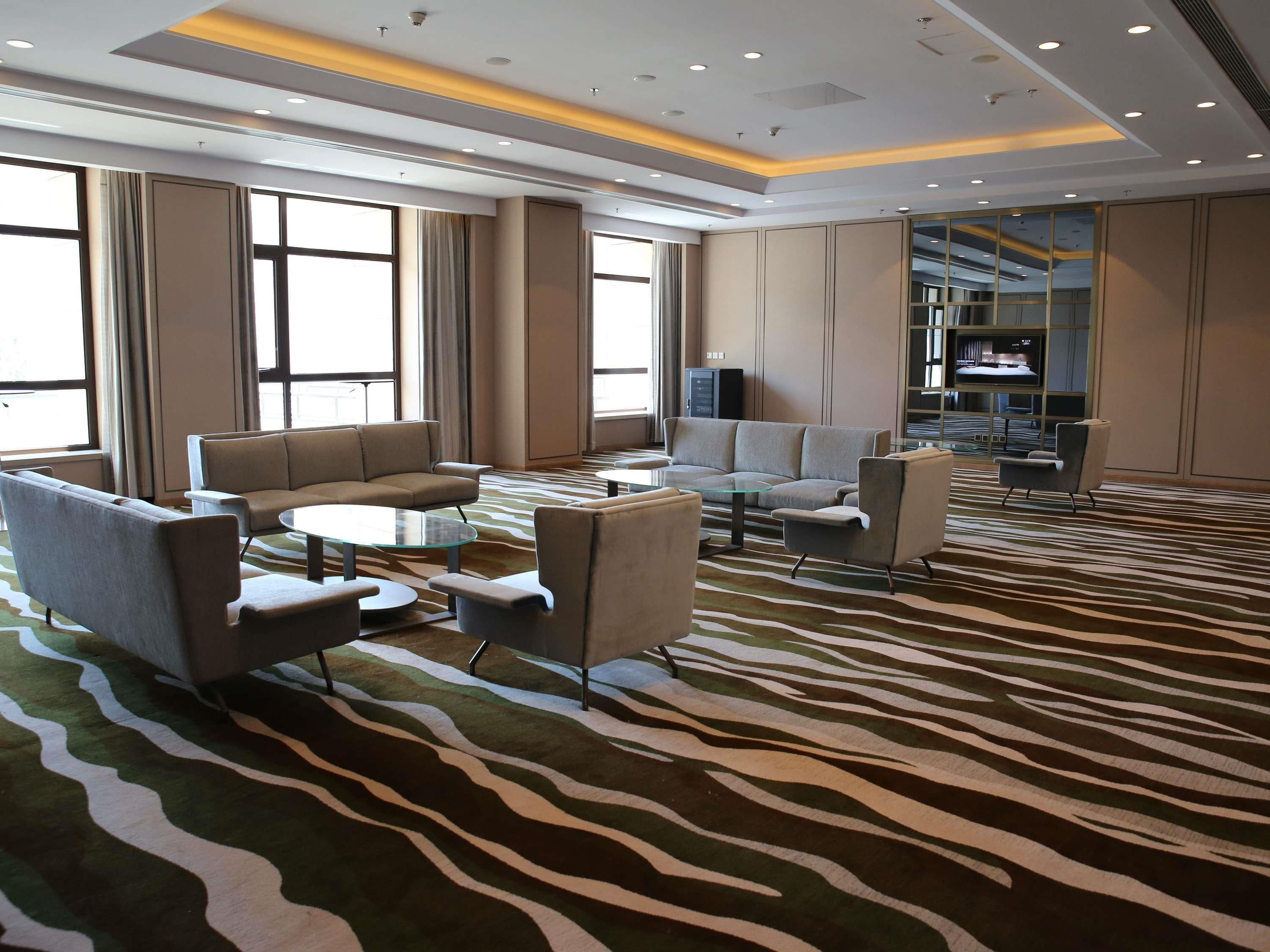 A modern hotel lobby with a zebra-patterned carpet, sofas, armchairs, a glass coffee table, and a television on a shelf.