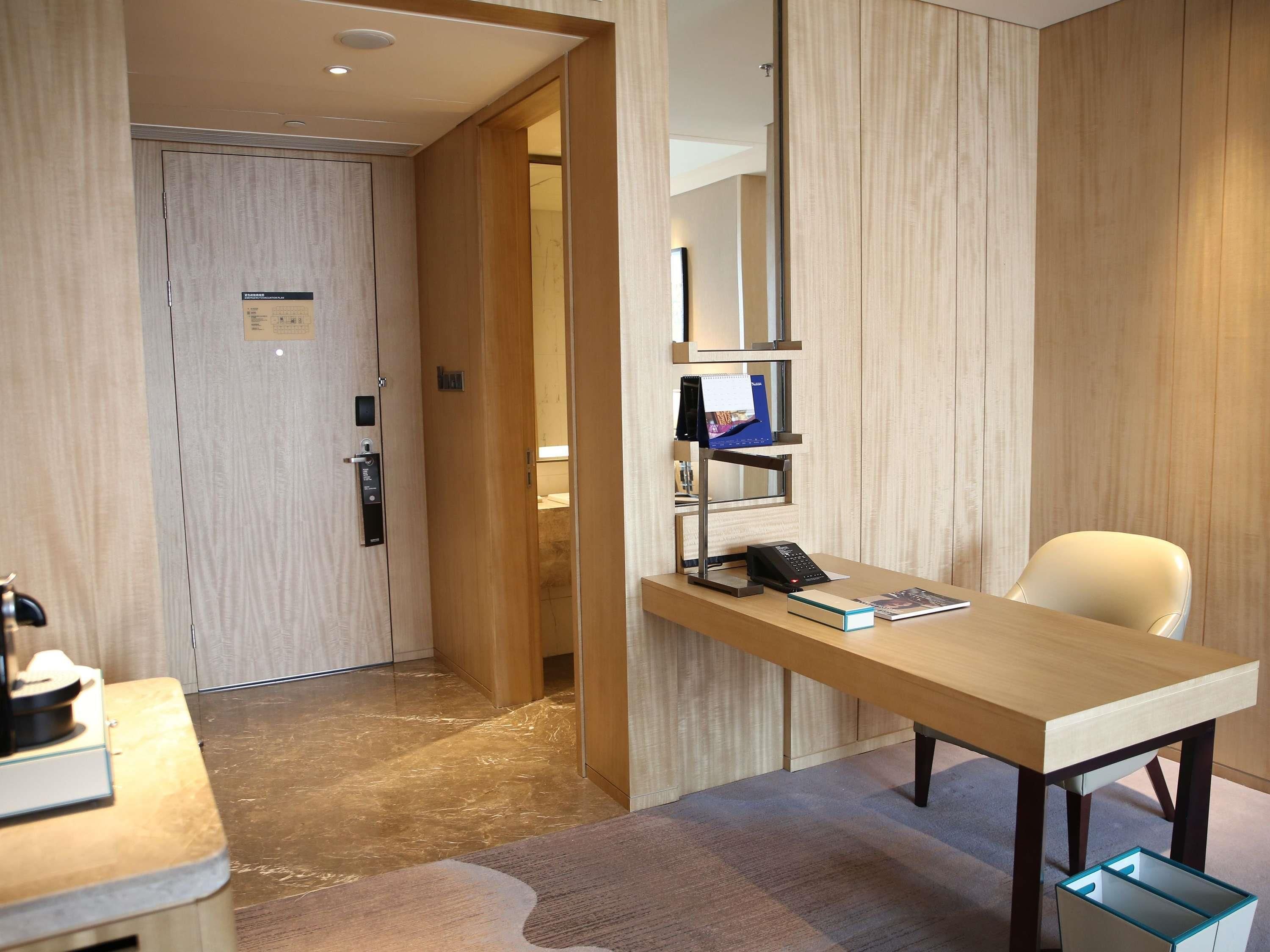 A hotel room with a wooden desk, a chair, a phone, a calendar, and a small shelf.