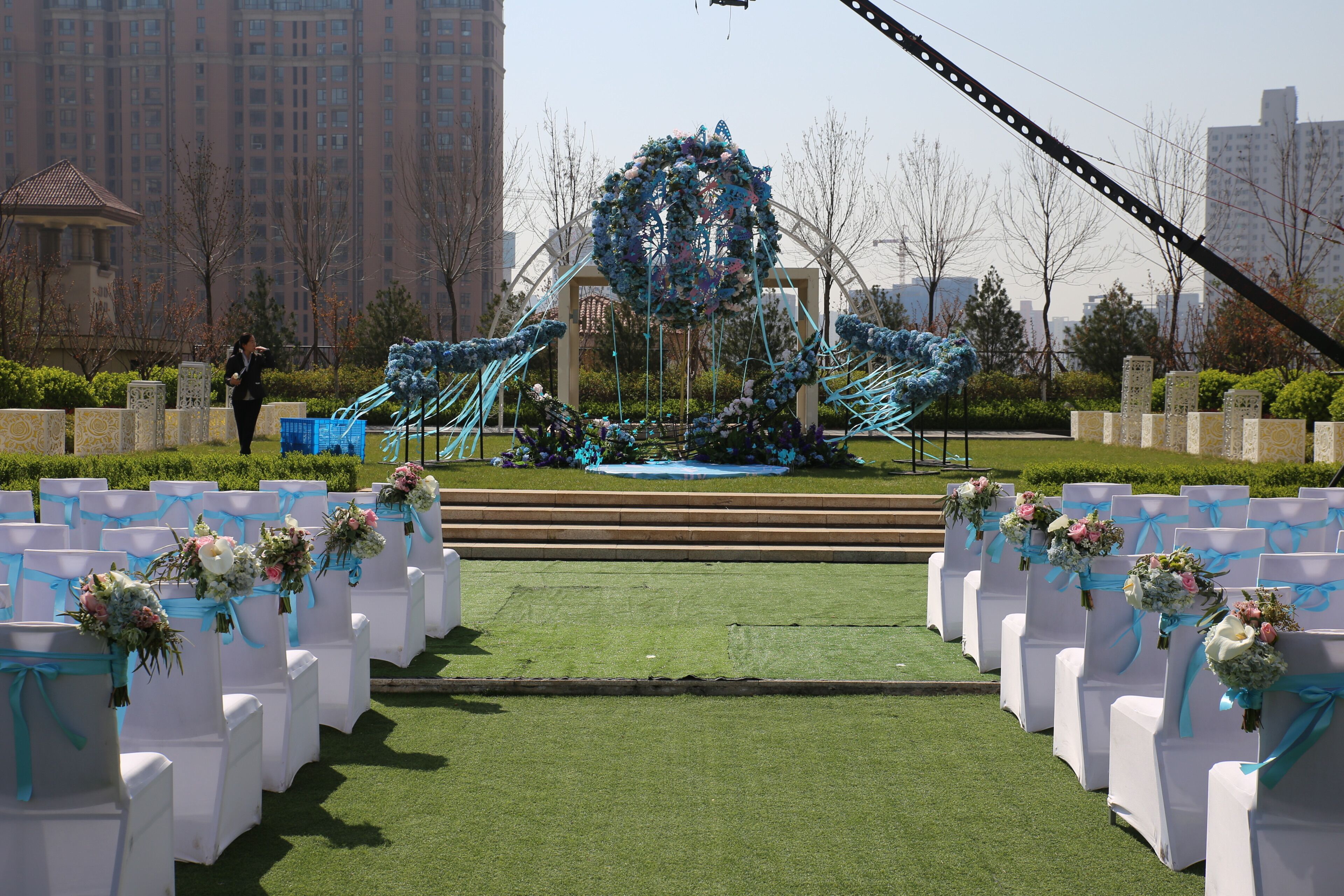 An outdoor venue with rows of white chairs, each adorned with floral arrangements and tied with blue ribbons.