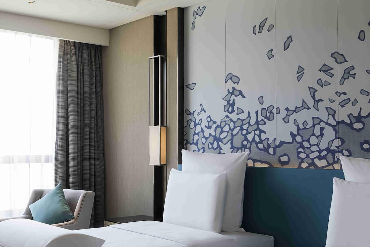 A hotel room with two beds, a large window with curtains, a wall with abstract art, and a tall, narrow light fixture.
