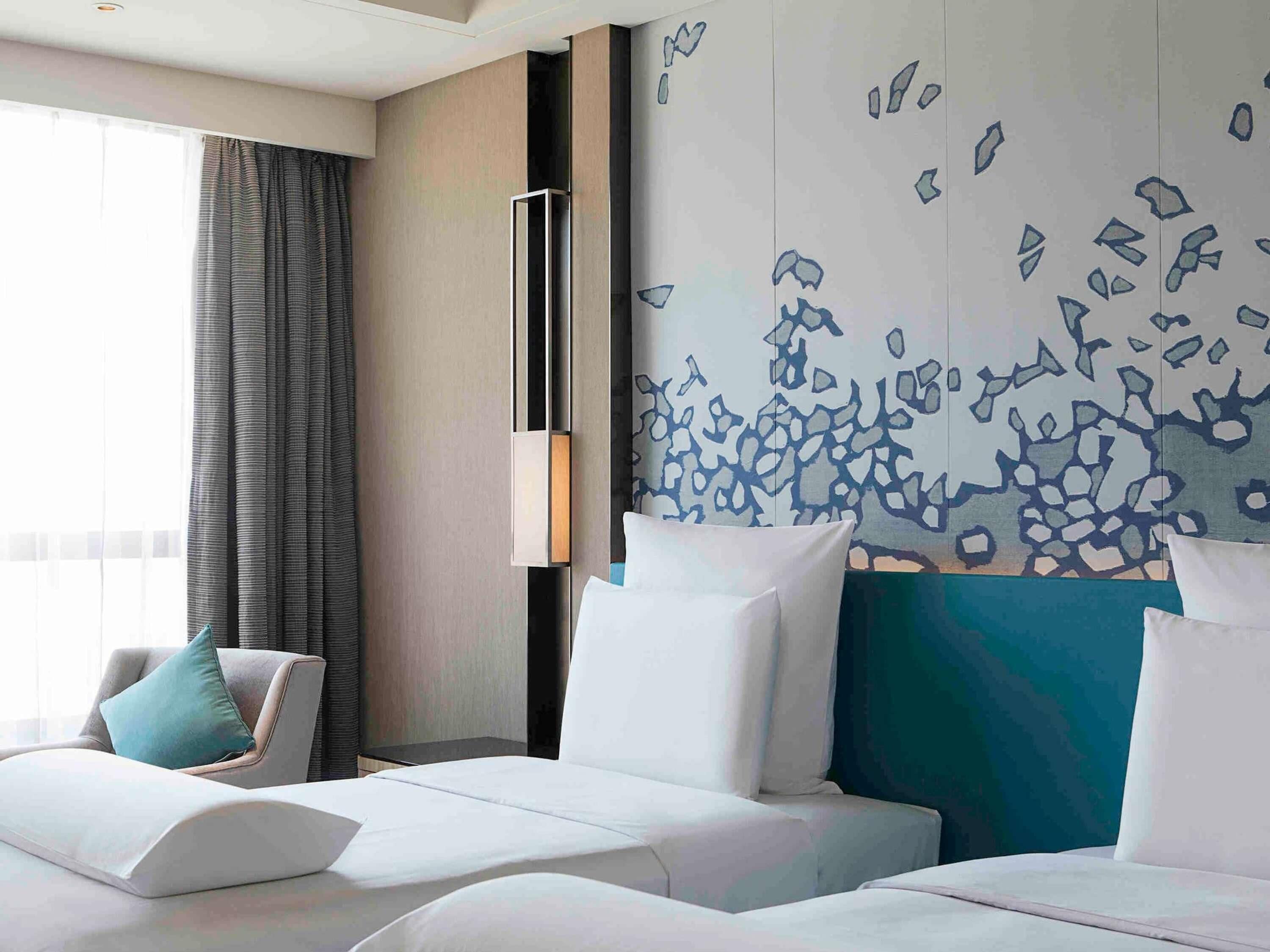 A hotel room with two beds, a large window with curtains, and a wall with abstract art.