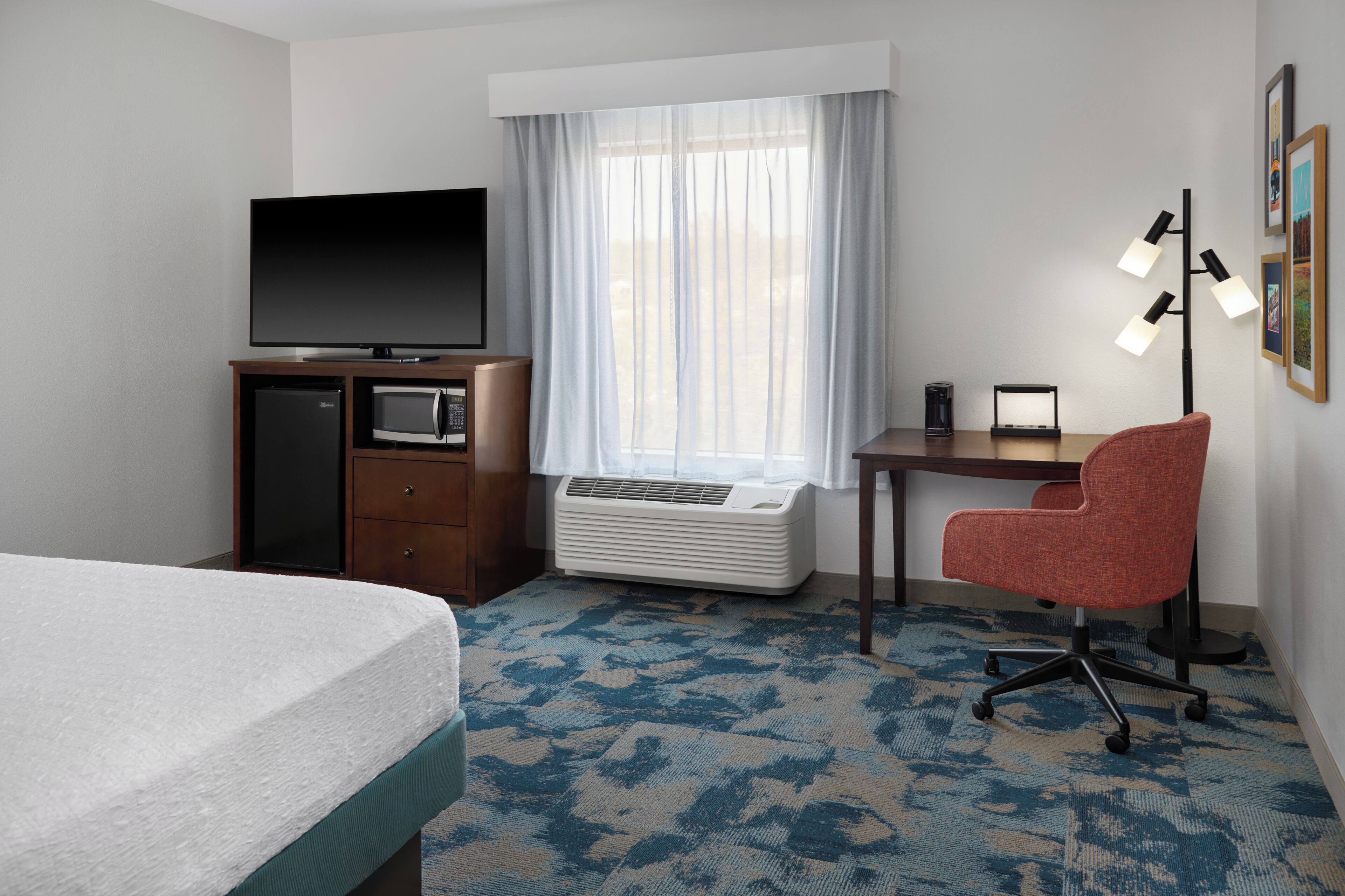 A hotel room with a bed, a desk with a chair, a television, a heater, and a window with curtains.