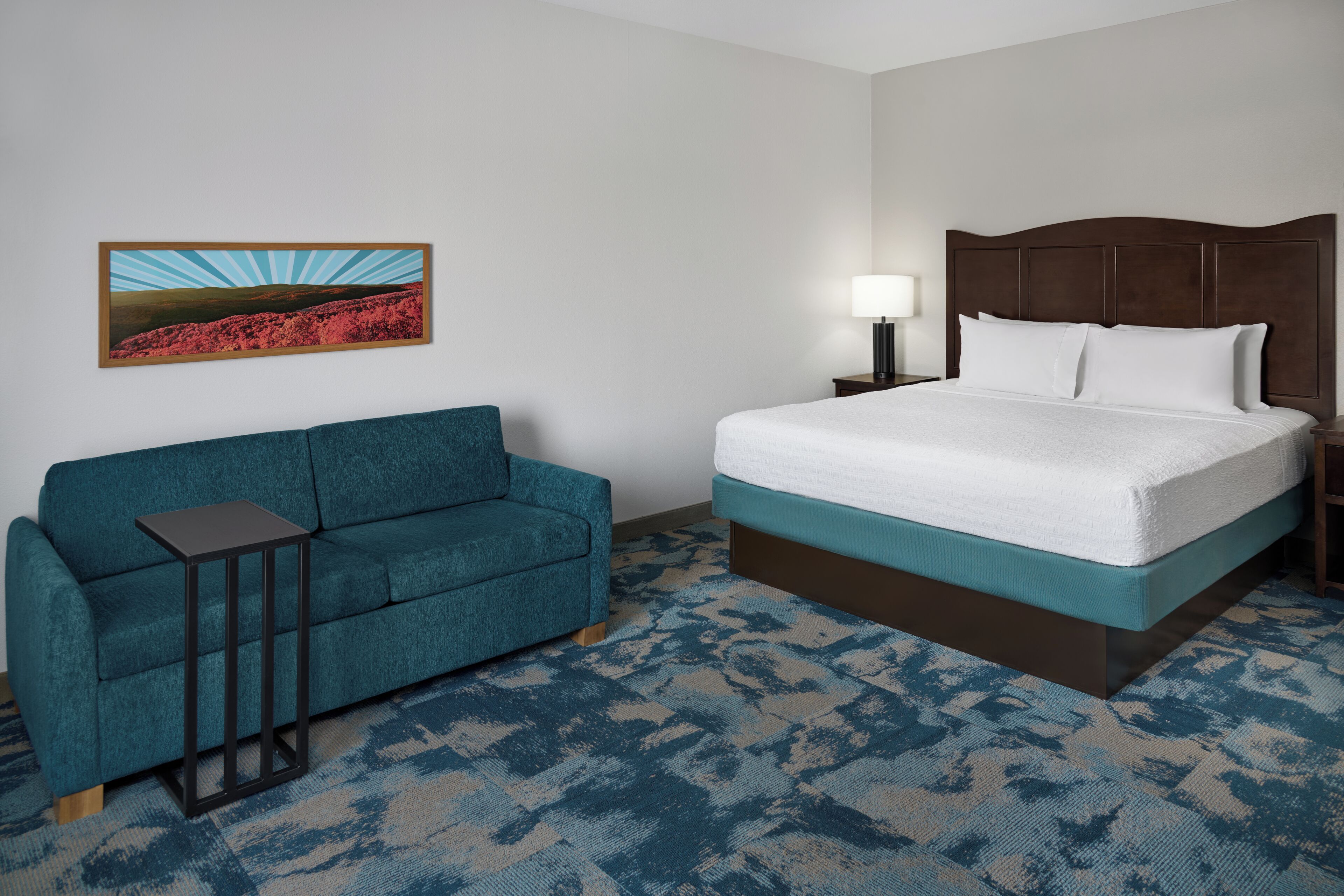 Una habitación de hotel con cama, sofá, mesita de noche, lámpara y un cuadro en la pared.