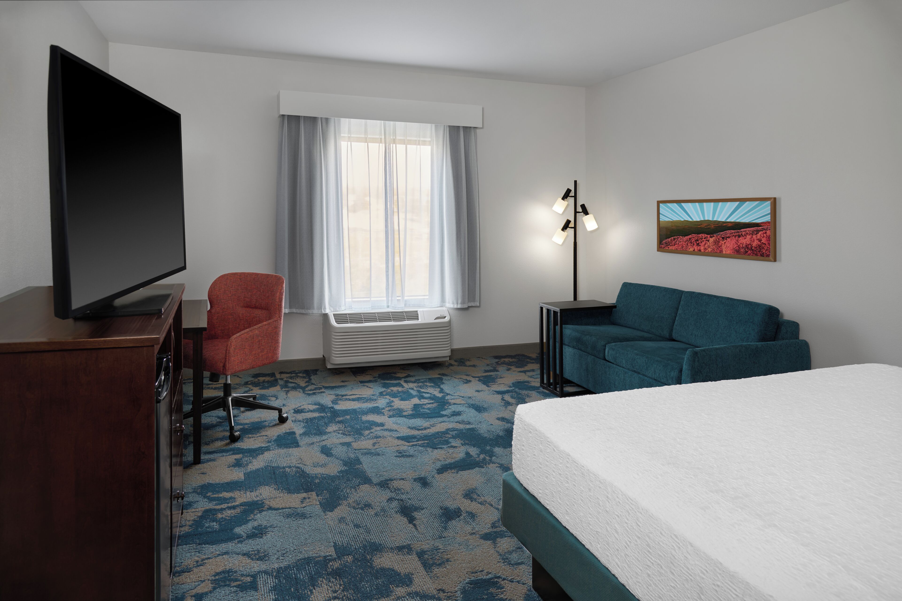 A hotel room with a bed, a TV, a red chair, a blue sofa, a window with curtains, and a wall-mounted lamp.