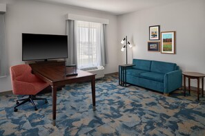 A hotel room with a flat-screen TV, a desk with a chair, a sofa, a standing lamp, and framed pictures on the wall.