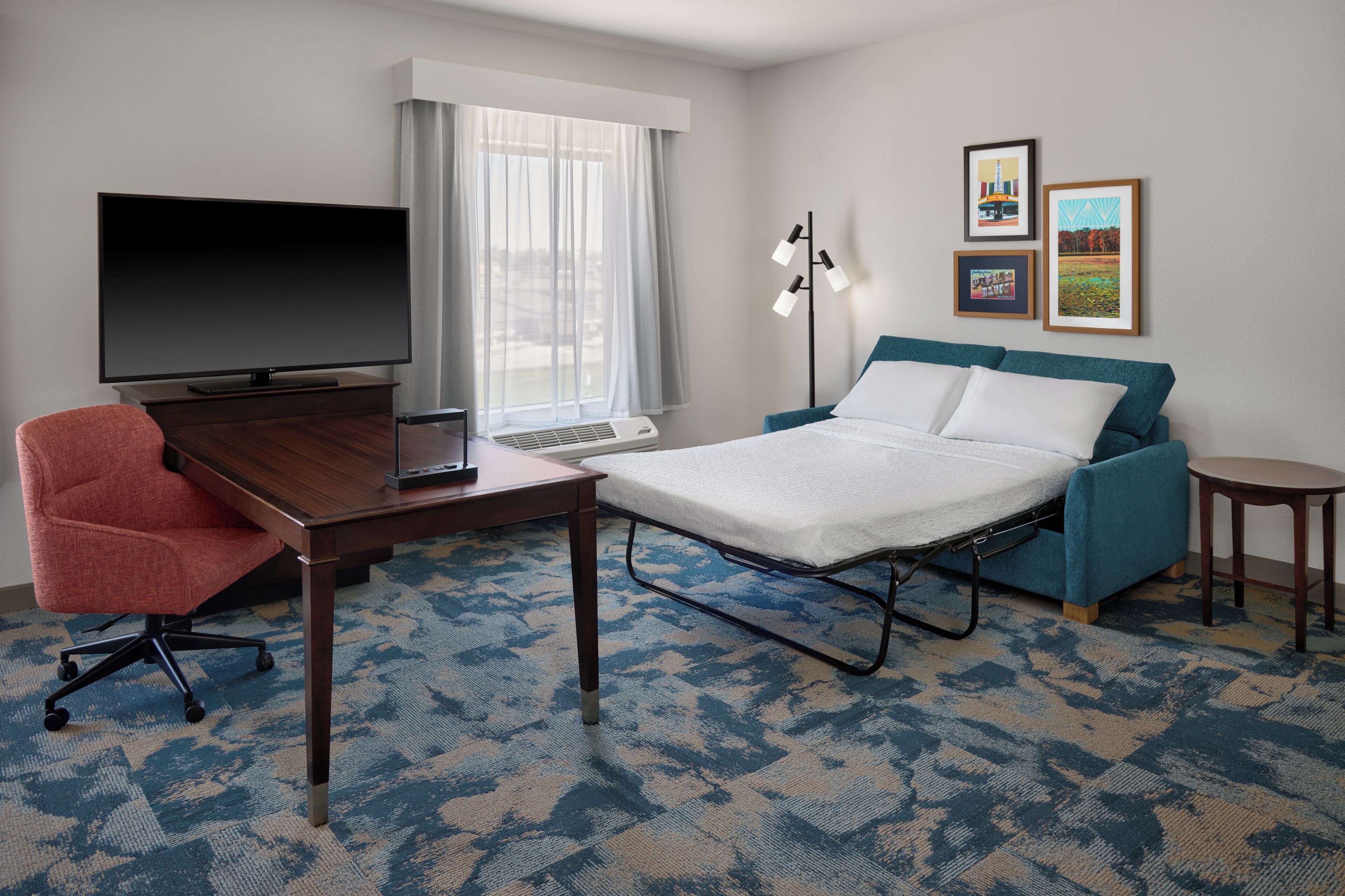 A hotel room with a bed, a desk with a television, a chair, a lamp, and framed pictures on the wall.