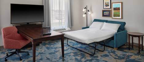 Une chambre d’hôtel avec un lit, un bureau avec une télévision, une chaise, une lampe et des tableaux encadrés au mur.