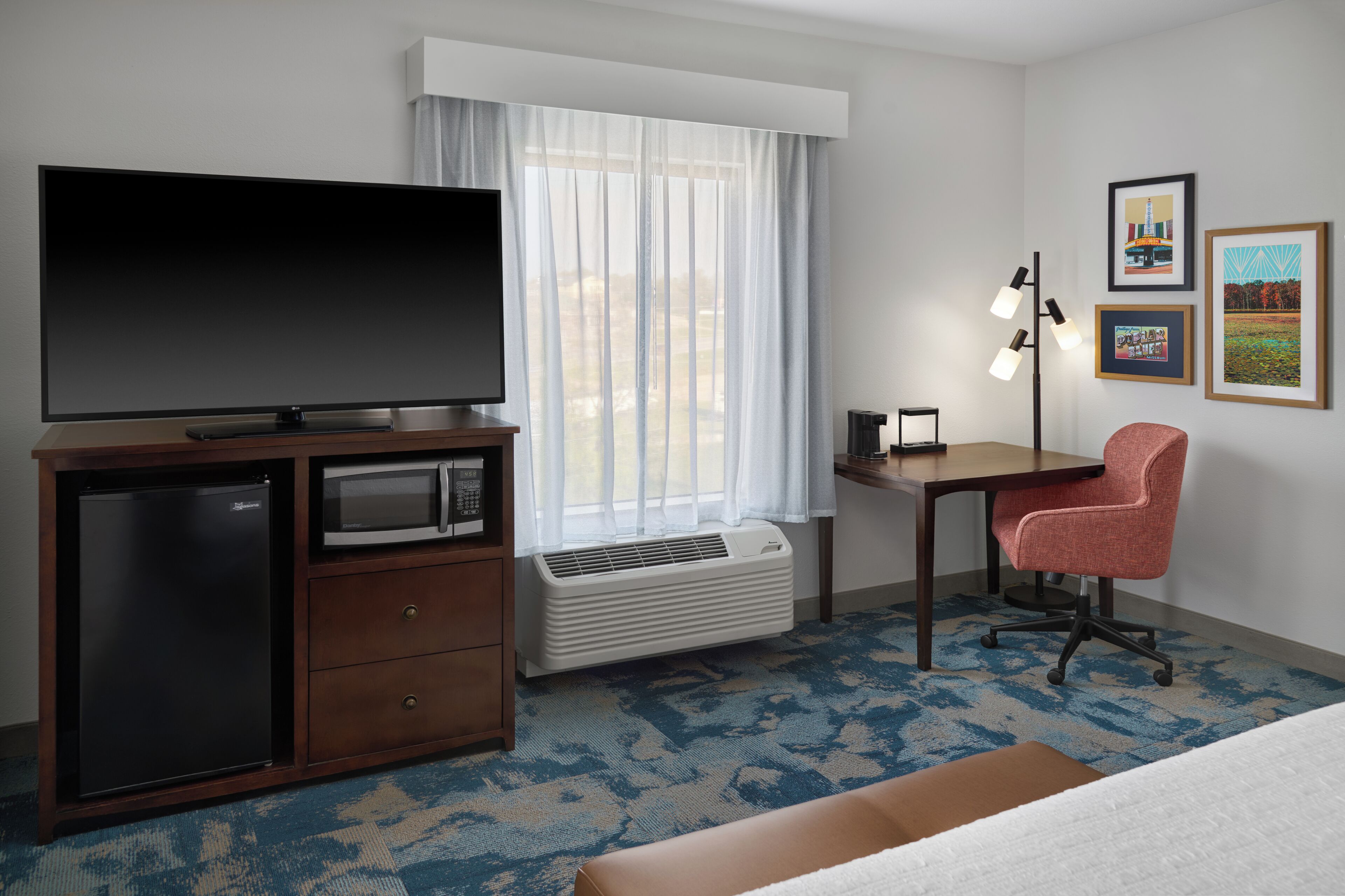 Ein Hotelzimmer mit Flachbildfernseher, Minikühlschrank, Mikrowelle, Schreibtisch, Stuhl, Stehlampe und gerahmten Bildern an der Wand.