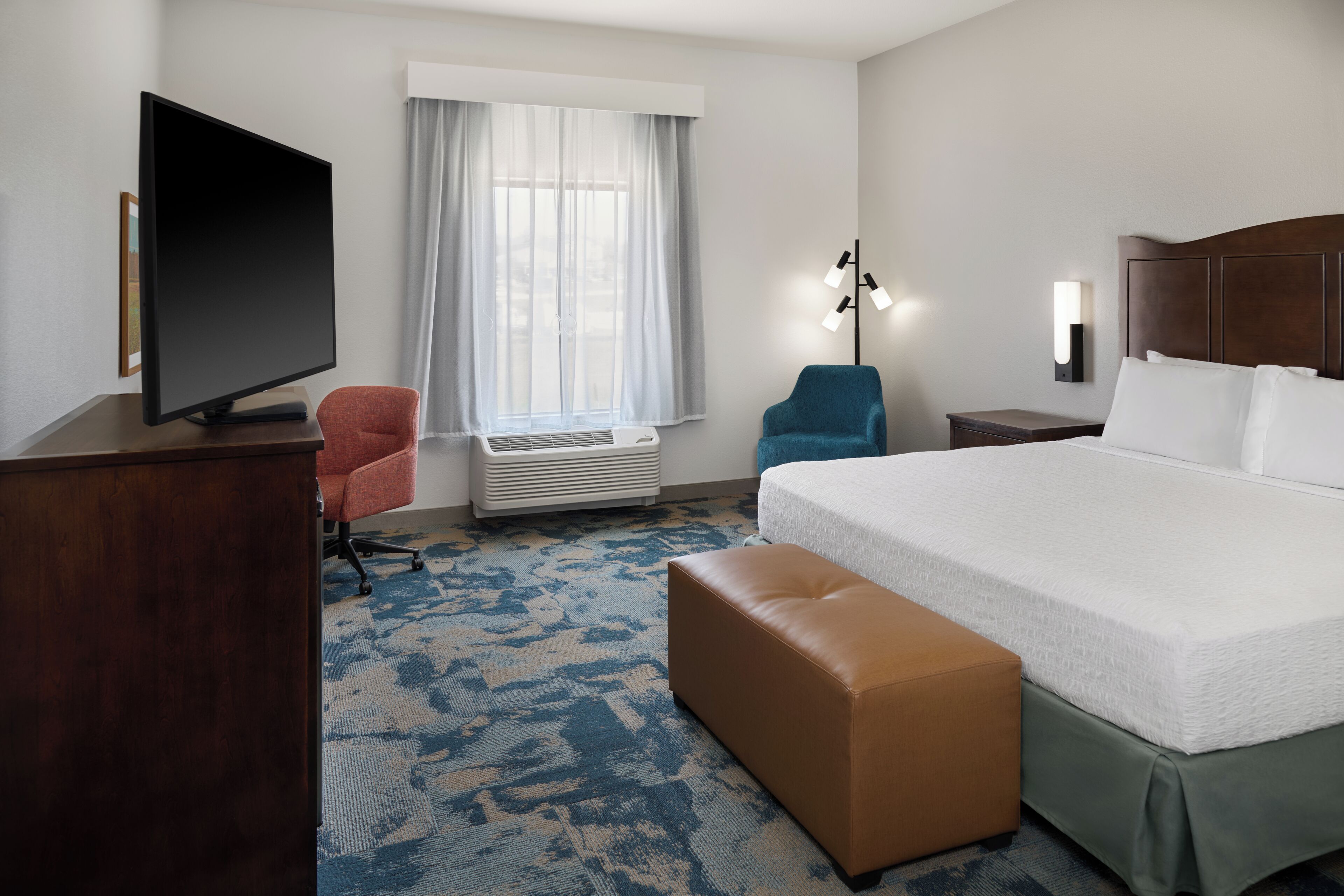 A hotel room with a bed, a television, a desk, a chair, a lamp, and a window with curtains.
