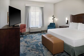 A hotel room with a bed, a television, a desk, a chair, a lamp, and a window with curtains.