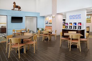A modern dining area with wooden tables and chairs, a mounted television, and a coffee station with various drink options.