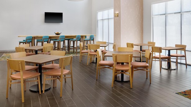 A spacious dining area with multiple tables and chairs, a flat-screen TV mounted on the wall, and large windows providing natural light.