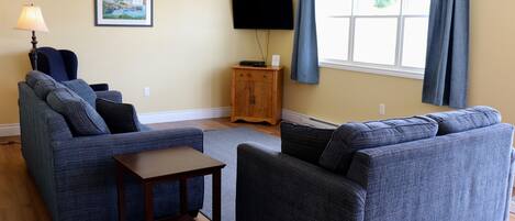 Una sala de estar con dos sofás, una mesita, un televisor sobre un mueble bajo y una ventana con cortinas.
