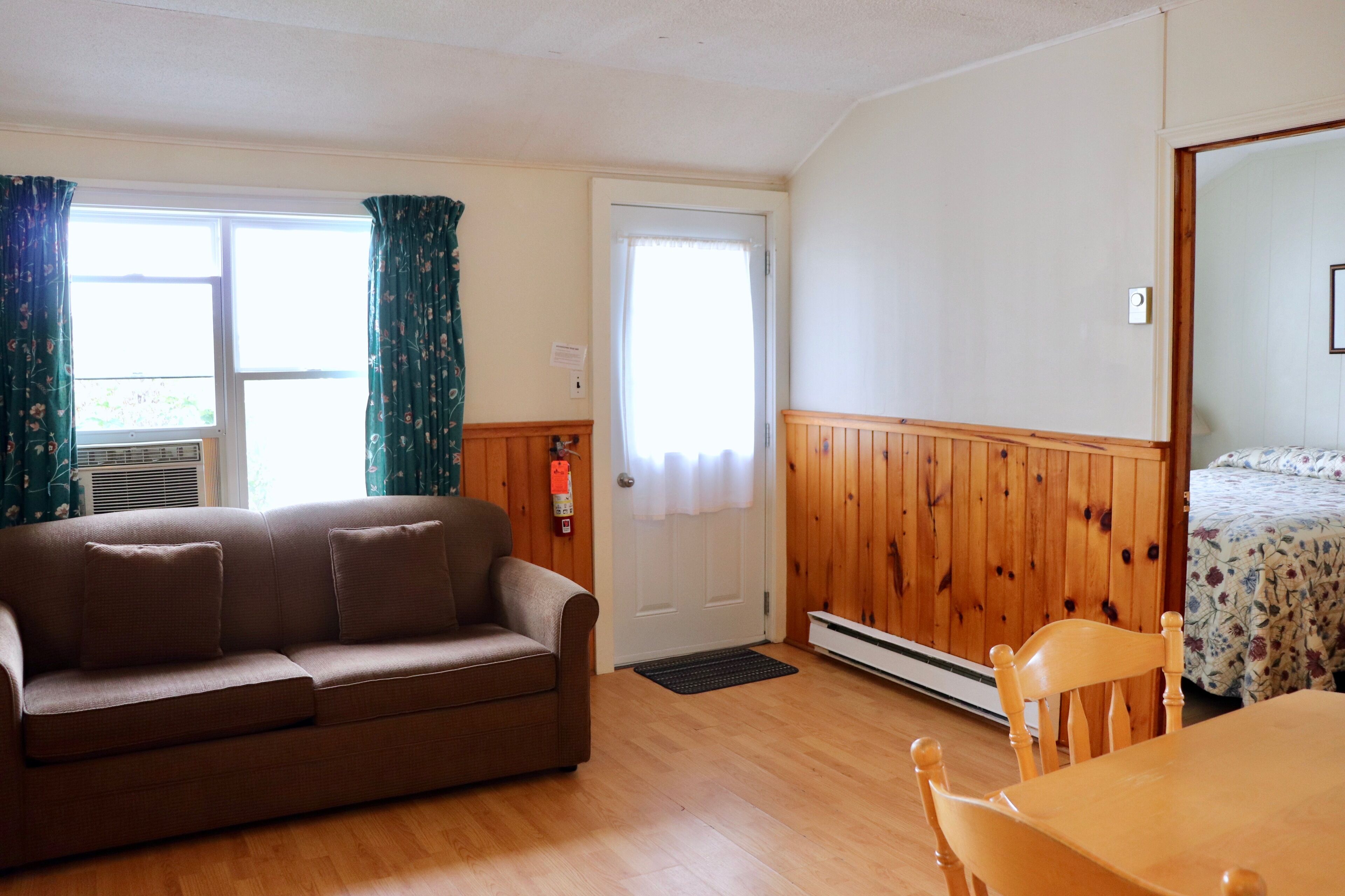Ein Zimmer mit Holzboden, einem braunen Sofa, einem Holztisch mit Stühlen, einem Bett mit Blumenmuster-Bettwäsche und einer Tür mit weißem Vorhang.