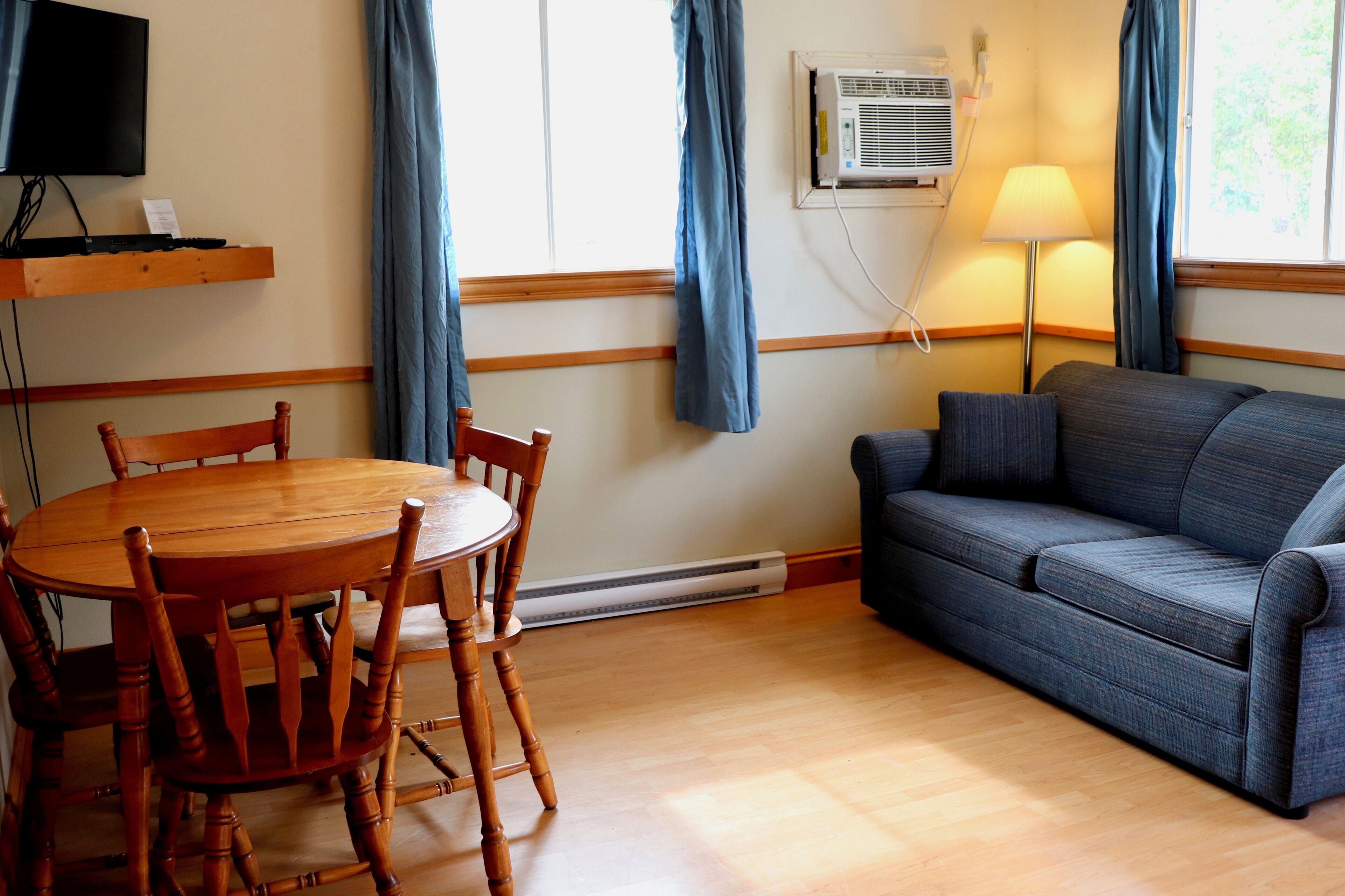 A room with a wooden dining table and chairs, a blue sofa, a TV on a shelf, and an air conditioning unit.