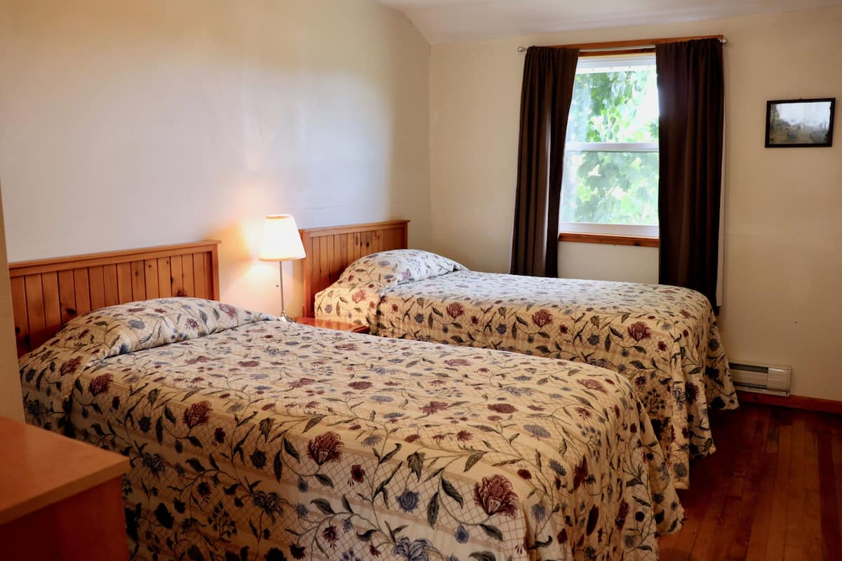Una habitación con dos camas, cabecera de madera, mesita de noche, lámpara, ventana con cortinas e imagen enmarcada en la pared.